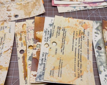 May include: A collection of rectangular library catalogue cards featuring printed bibliographic text and circular punched holes. The off-white paper cards are stained with various shades of brown, orange, and yellow ink splatters and blotches.