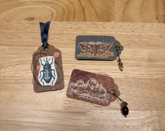 May include: Three rectangular paper tags featuring nature-themed illustrations. One tag displays a blue beetle with a dark ribbon, another shows a moth on a blue background with beads, and the third features a mushroom design with dark beads.