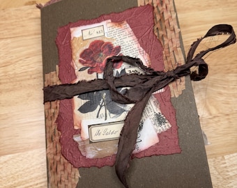 May include: A handcrafted journal with a dark brown cover and a burgundy accent. The journal features a floral design and is tied with a dark brown ribbon. The journal has the text "No 813" and "Me Galdo" on the front.