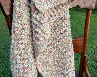 May include: A hand-knitted scarf in a speckled pattern of beige, blue, yellow, and pink. The scarf is draped over the back of a wooden chair. The yarn appears soft and textured, suggesting a cosy accessory for cooler weather.