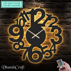 Op de afbeelding: Een zwarte klok met verlichte cijfers in een cirkelvormig ontwerp. De klok heeft een afstandsbediening met meerdere gekleurde LED-lampjes. De tekst "LED light color can be changed" is zichtbaar in de linkerbovenhoek.