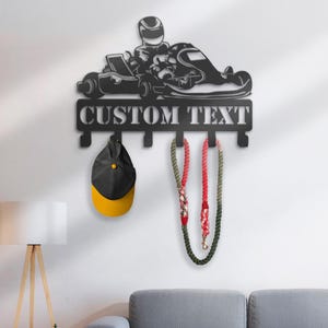 May include: Black metal wall-mounted coat rack shaped like a go-kart with a driver. The rack has five hooks and the text "CUSTOM TEXT" is printed on the top. A black and yellow baseball cap and two red and green ropes are hanging on the hooks.