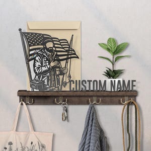 May include: A wooden wall shelf with hooks, displaying a metal cutout of a person with an American flag. The shelf also holds a small plant in a white pot and the words "CUSTOM NAME". A tote bag, keys, and a scarf hang below.