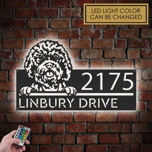 Può includere: Un cartello per numeri civici in metallo nero con una silhouette di un barboncino bianco che sporge dalla parte superiore. Il cartello è illuminato da luci LED bianche che illuminano il numero civico "2175" e il nome della strada "LINBURY DRIVE". Il cartello è montato su un muro di mattoni.