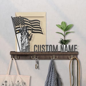 May include: A wooden wall shelf with hooks, a plant, and a metal sign that reads "CUSTOM NAME." The shelf also features a cutout of the Statue of Liberty holding a flag. A tote bag, keys, and a scarf hang from the hooks.