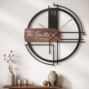May include: A circular wall clock featuring a black metal frame with geometric lines and a horizontal wooden panel. The panel displays the white text The Smiths and EST. 2024. The clock has copper-colored hands and the number 12 at the top.