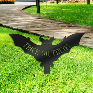 May include: Black metal silhouette of a bat with the words "Trick or Treat" cut out. The bat is on a green lawn.