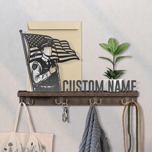 May include: A wooden wall shelf with hooks, a decorative metal cutout of a police officer saluting the American flag, and the text "CUSTOM NAME". A small potted plant and a tote bag are also visible.