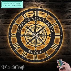 May include: Black metal clock with Roman numerals and a compass design. The clock has a light that can be changed to different colors using a remote control. The clock also has the text "NEVADA" and coordinates "38°53'13.5"N 115°47'30.5"W" on it.