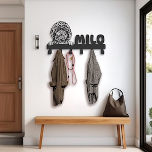 May include: Black metal coat rack with a dog silhouette and the name "Milo" on it. The rack has four hooks and is mounted on a white wall. There is a brown leather bag hanging on one of the hooks and two jackets hanging on the other hooks. A red and white leash is hanging on one of the hooks.