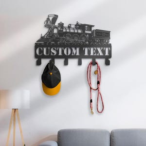 May include: Black metal wall-mounted coat rack featuring a train design with the text "CUSTOM TEXT". A black and yellow baseball cap and a red and white dog leash hang from the hooks.