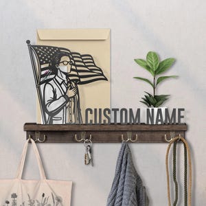 May include: A wooden shelf with hooks, holding a tote bag, keys, and a scarf. Above, a metal cutout of a person with an American flag, and the words "CUSTOM NAME". A small potted plant sits to the right.