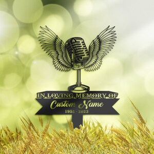 May include: Black metal memorial sign featuring a microphone with wings, above a banner that reads "IN LOVING MEMORY OF" and space for a custom name, with the years 1955-2023. The sign is set against a blurred green and yellow background.