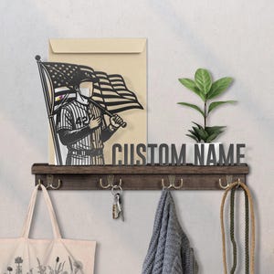 May include: A wooden shelf with hooks, holding a tote bag, keys, and a scarf. Above, a metal cutout of a baseball player holding an American flag, and the words "CUSTOM NAME". A small plant in a white pot sits on the shelf.