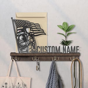 May include: A wooden wall shelf with hooks, a metal cutout of a firefighter holding an American flag, and the text "CUSTOM NAME". A tote bag, keys, and a scarf hang from the hooks. A small plant in a white pot sits on the shelf.