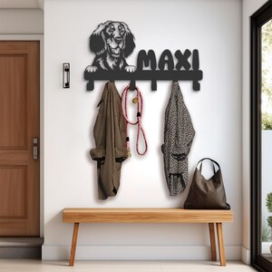 May include: A black metal wall-mounted coat rack featuring a dog silhouette and the name "MAXI". The rack holds coats, a leash, and a tote bag. A wooden bench sits below the rack. The wall is white, and a wooden door is visible.