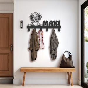 May include: Black metal coat rack with four hooks and a dog silhouette. The rack has the name "Max" written on it. Two coats and a leash are hanging on the hooks. The rack is mounted on a white wall above a wooden bench.