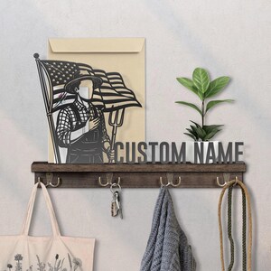 May include: A wooden shelf with hooks, holding a tote bag, keys, and a scarf. Above, a metal sign reads "CUSTOM NAME." A cutout depicts a farmer with an American flag and a pitchfork. A small plant in a white pot sits on the shelf.