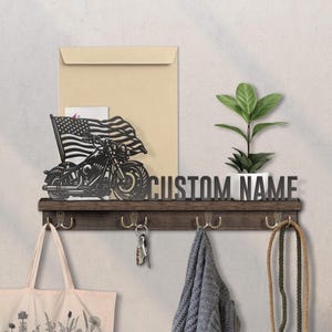 May include: A dark wood wall-mounted shelf featuring a black metal cutout of a motorcycle with an American flag, the text CUSTOM NAME, and four metal hooks. A small green potted plant sits on the shelf next to a beige envelope.