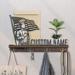 May include: A wooden shelf with hooks, holding a tote bag, keys, and a scarf. Above, a metal sign reads "CUSTOM NAME." A cutout depicts a chef saluting the American flag, with a plant in a white pot.