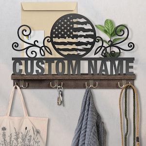 May include: A black metal wall-mounted organizer with the text "CUSTOM NAME" and an American flag design. It features hooks for keys, coats, and bags, and a small shelf. A tote bag, keys, and a scarf hang from the hooks.