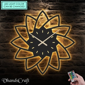 May include: A black clock with a geometric design and LED lights that can be changed with a remote control. The clock has a black face with white hands and a black frame. The LED lights are illuminating the geometric design, which is made of black metal. The remote control has a variety of colors that can be selected.