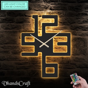 May include: Black modern wall clock with illuminated numbers and a remote control to change the LED light color. The clock is mounted on a brick wall. "LED light color can be changed" is written on a turquoise rectangle at the top of the image.