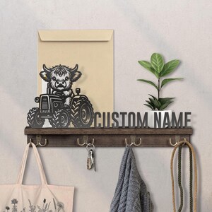 May include: A dark wood wall-mounted shelf with four metal hooks, featuring a black cutout of a Highland cow driving a tractor on the left side. The shelf includes the text CUSTOM NAME in black lettering and holds a small green potted plant.
