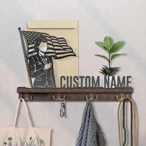 May include: A wooden shelf with hooks, holding a tote bag, keys, and a scarf. Above, a metal sign reads "CUSTOM NAME" and a plant sits in a white pot. A black and white illustration of a person saluting the American flag is displayed.