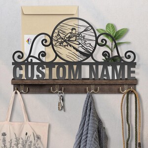 May include: A personalized wall-mounted key holder with a dark brown wooden base and metal hooks. The design features a kayaker in a circular frame, with the text "CUSTOM NAME" below. A tote bag, keys, and a scarf hang from the hooks.