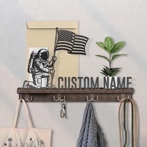 May include: A dark brown wall shelf with hooks, a metal astronaut figure holding an American flag, and the text "CUSTOM NAME". A small plant in a white pot sits on the shelf. A tote bag, keys, and a scarf hang below.