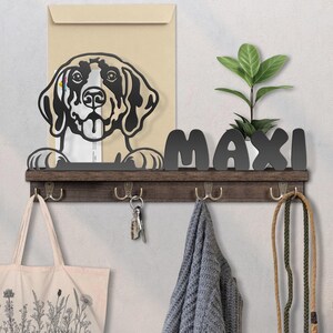 May include: A wooden wall-mounted dog leash and key holder with the name "MAXI" in black letters. The holder features a black metal dog silhouette, hooks, and a small plant. A tote bag, keys, and a leash hang from the hooks.