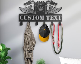 Könnte beinhalten: Schwarzer Wand-Kleiderhaken aus Metall mit einem Motorrad-Design und dem Text "CUSTOM TEXT". Der Haken hat fünf Haken und ist mit Flügeln verziert. Ein Hut, eine Jacke und zwei Leinen hängen an den Haken.