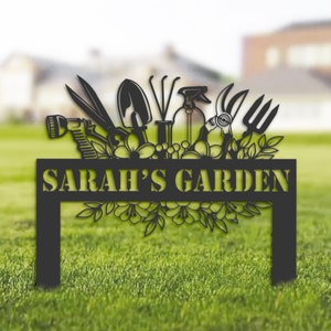 Puede incluir: Señal de jardín de metal negro con un diseño floral y herramientas de jardinería. El letrero dice "SARAH'S GARDEN".