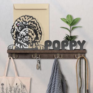 Könnte beinhalten: Ein Wandregal aus Holz mit Haken, eine Metall-Hunde-Silhouette und der Name "POPPY" in grauen Buchstaben. Eine Tragetasche, Schlüssel und eine Leine hängen an den Haken. Eine kleine Pflanze steht auf dem Regal.