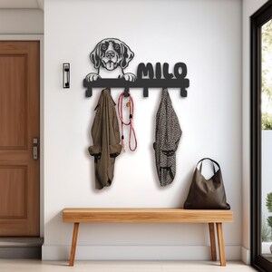 May include: Black metal coat rack with a dog silhouette and the name "Milo" above four hooks. The rack is mounted on a white wall above a wooden bench. Two coats and a leash are hanging on the hooks.