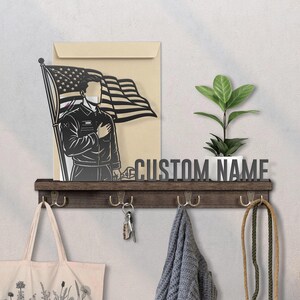 May include: A dark wooden wall shelf with hooks, holding a tote bag, keys, and a scarf. It features a cutout of a person saluting the American flag, a plant, and the text "CUSTOM NAME".