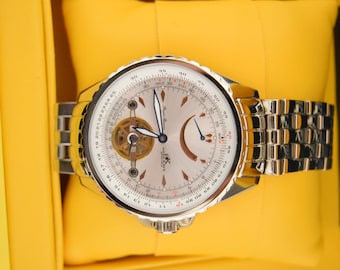 May include: A silver wristwatch with a white face and a silver band. The watch has a skeletonized movement and a date window at the 3 o'clock position. The watch is in a yellow box.