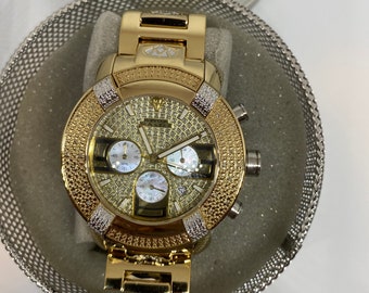 May include: A gold-tone watch with a diamond-encrusted bezel and a white face. The watch has a chronograph function and a date window. The watch is in a white box with a silver mesh pattern.