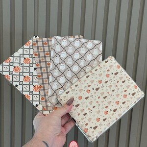 Puede incluir: Una colección de fundas para teléfonos con temática de Halloween y varios diseños. Los diseños incluyen fantasmas, calabazas y motivos de calaveras en naranja, blanco y marrón sobre un fondo neutro. Las fundas se muestran contra una pared gris texturizada.