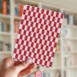 May include: A tablet case featuring a red and pale pink chequered design, embellished with diamond motifs. The case is held up against a blurred backdrop of a bookshelf.