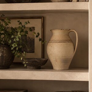 May include: A beige ceramic pitcher with a textured, matte finish and a single handle. The pitcher features two thin, horizontal brown bands encircling the upper body. It has a wide, flared rim and a tapered base, standing on a shelf.
