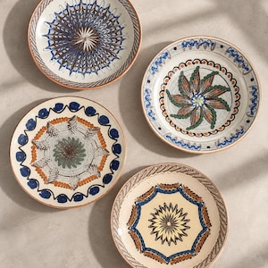 May include: Four round ceramic plates with cream-colored bases and hand-painted designs. Each plate features distinct patterns in blue, brown, green, and orange, including radial starbursts, floral motifs, and geometric borders along the edges.