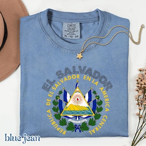 A blue t-shirt with a graphic design of the Salvadoran flag, along with a necklace and a hat, all placed on a white surface.