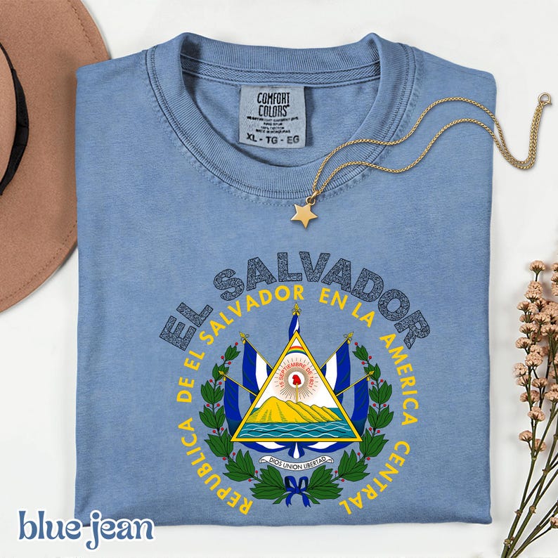 A blue t-shirt with a graphic design of the Salvadoran flag, along with a necklace and a hat, all placed on a white surface.