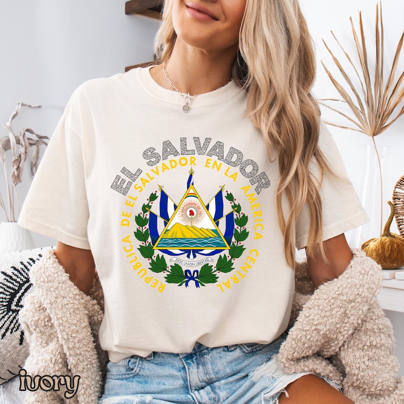 A woman wearing a white t-shirt with the text "EL SALVADOR EN LA AMERICA" printed on it, along with a graphic of the national coat of arms of El Salvador.