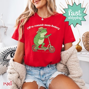 以下が含まれることがあります： 「Off to commit mom foolery」の文字と、スクーターに乗った漫画の蛙が描かれた赤いTシャツ。 蛙は緑色で帽子をかぶっています。 シャツには「速い発送」のグラフィックも付いています。