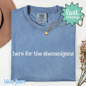 以下が含まれることがあります： 「here for the shenanigans」の白文字が入った、ブルージーンズ色のTシャツ。金の星のペンダントが金のチェーンからぶら下がっています。シャツは折りたたまれており、「Comfort Colors」と書かれたタグが付いています。
