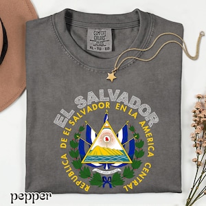 A gray t-shirt with a graphic design featuring the coat of arms of El Salvador, along with the text "EL SALVADOR EN LA AMERICA CENTRAL REPUBLICA".