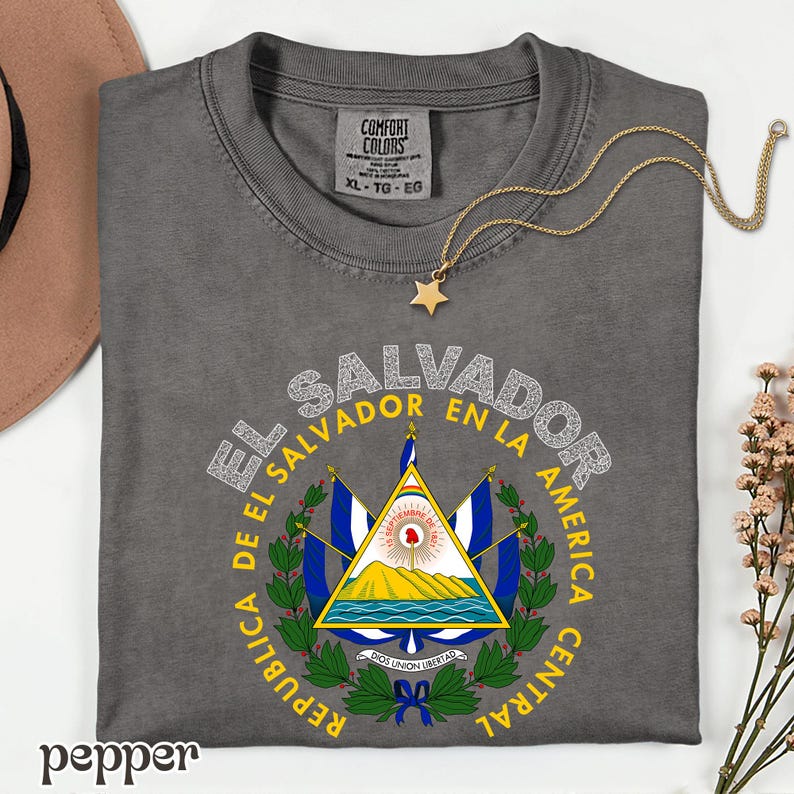 A gray t-shirt with a graphic design featuring the coat of arms of El Salvador, along with the text "EL SALVADOR EN LA AMERICA CENTRAL REPUBLICA".